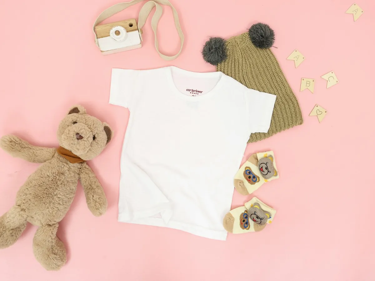 White kids t-shirt flat-lay with a teddy bear, knit beanie, and baby socks on a soft pink background